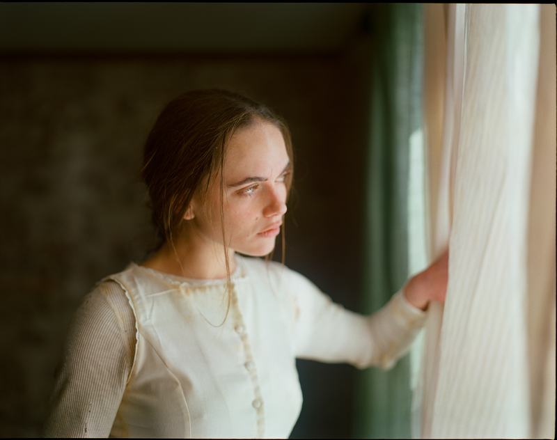 A sister in white parts a curtain and looks out the window.