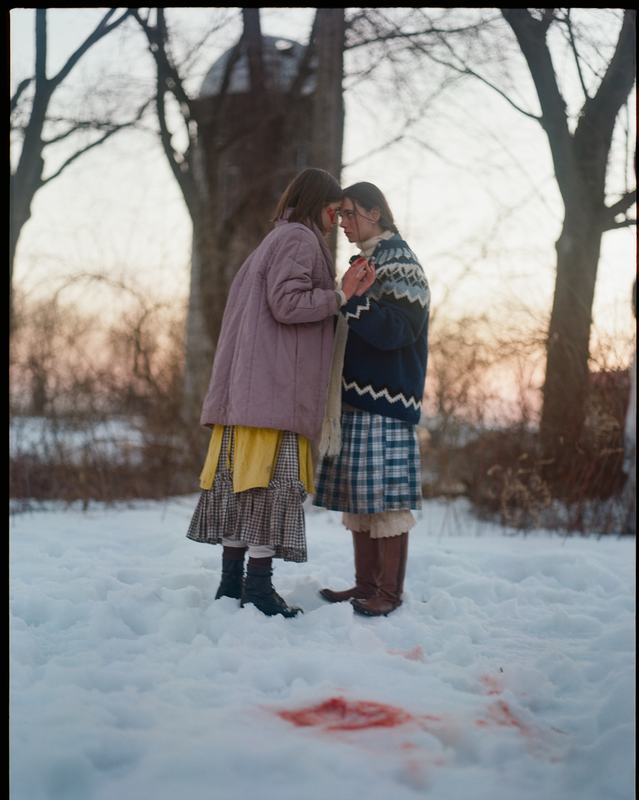 The sisters stand close in the snow; a red stain spreads at their feet.