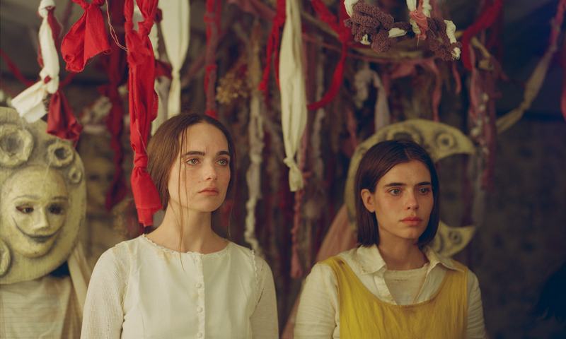 The sisters in convent dress stand beneath hanging ribbons beside a masked figure.