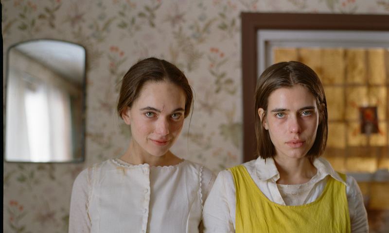 The sisters, one in a yellow pinafore, look straight into the lens in a papered room.