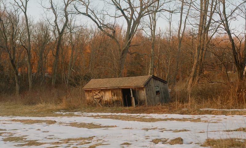 A weathered cabin at the edge of a thawing, snow-covered field.