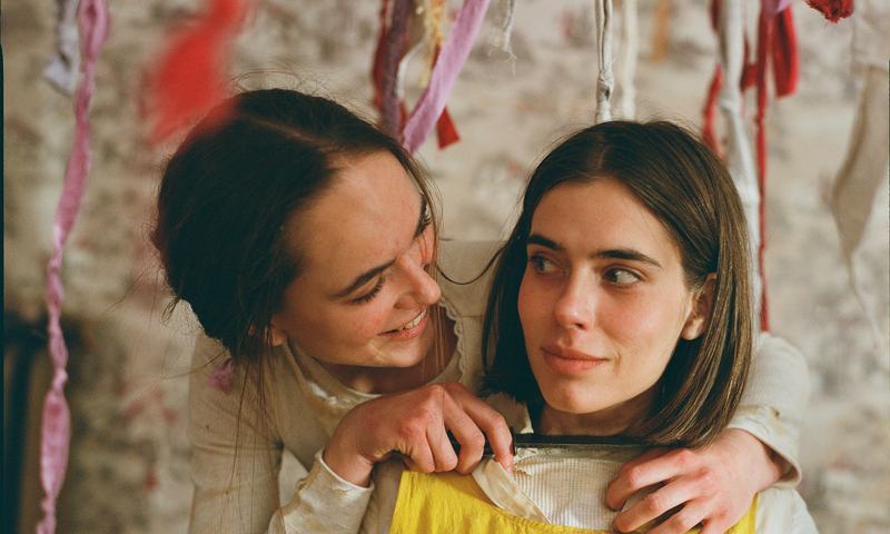 One sister holds the other from behind, her chin on her shoulder.