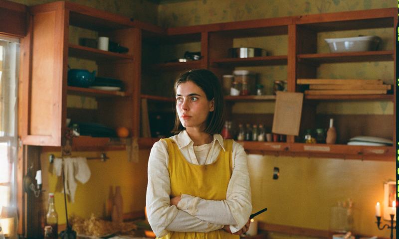 A sister in a yellow apron stands with folded arms in a candlelit kitchen.