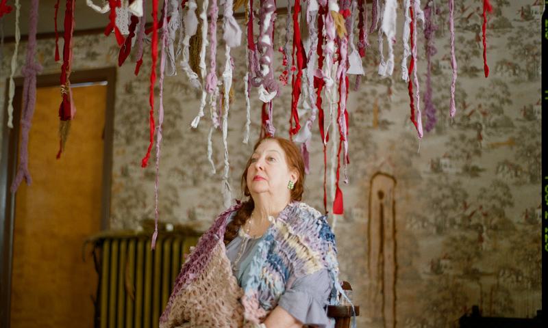 A matron in a knitted shawl sits beneath hanging ribbons, looking up.
