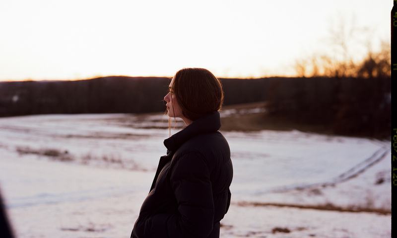A figure in a dark coat stands in the snow at sunset.